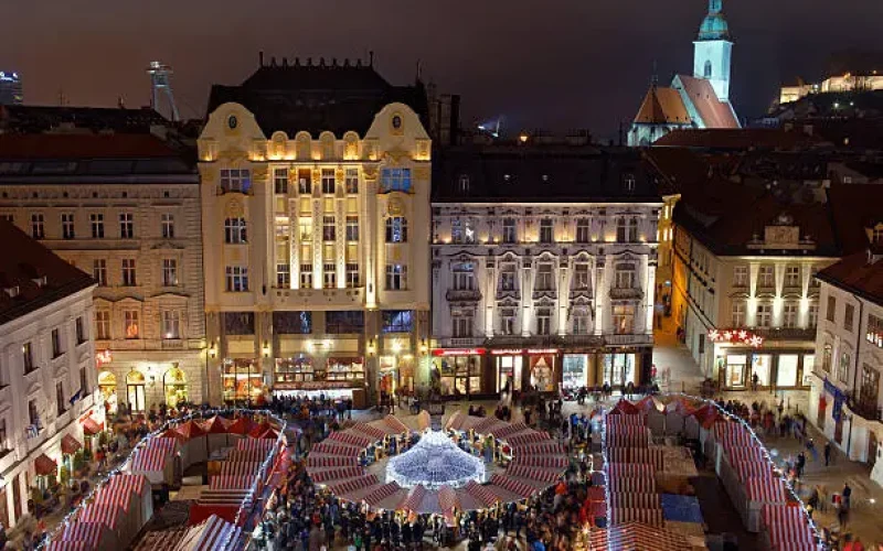 Drohnenbild von einem weihnachtlich geschmückten und beleuchteten historischen Hauptplatz mit beleuchtetem Kirchturm im Hintergrund und vielen Besuchern im Vordergrund bei Nacht