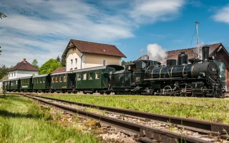 Frühlingslandschaft mit Dampfzug vor Bahnhofsgebäude stehend, Geleise im Bildvordergrund, blauer Himmel mit weißen Wolken