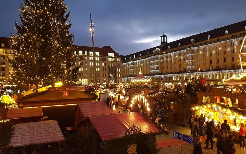 Großer Innenhof mehrstöckiger Gebäude mit dekorierten Holzkütten, einem großen Tannenbaum am linken Bildrand, alles in festlicher Weihnachtsbeleuchtung, Menschenansammlung bei den Hütten im Bildvordergrund