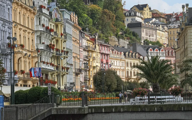 Bunte Häuserfronten entlang einer Stadtpromenade, mehrstöckige Hauser auf dem Hügeligen Gelände in der rechten Bildecke, entlang der unteren Bildseite ein Brückengeländer, hinter dem sich bunte Blumenbeete, grüne Büsche, Palmen und eine Baumallee zwischen den Häuserfronten entlang winden