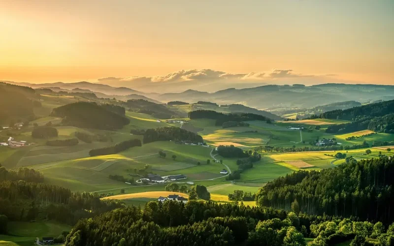 Eine Luftaufnahme von sanften, grünen Tälern und Hügeln mit kleinen Waldstücken und vereinzelten Bauernhöfen dazwischen, am unteren Bildrand bewaldeter Hügel, am Horizont Hügelketten im Dunst des orangefarbenen Sonnenuntergangs, eine Wolkendecke schwebt über den Hügelspitzen