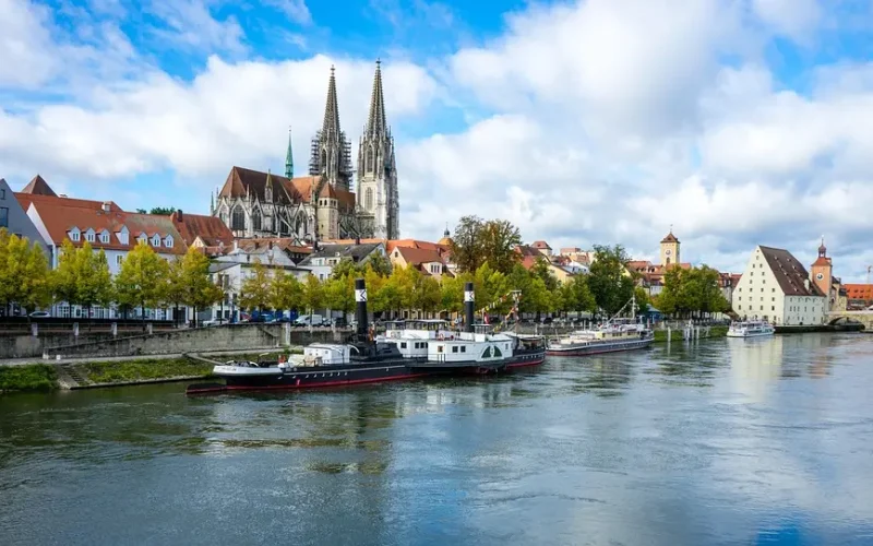 Blick von einem Fluss auf einen Kai, die dahinterliegende Stadt und einem Dom mit zwei Türmen und Brückenbögen, drei am Kai angelegte Schiffe im Vordergrund