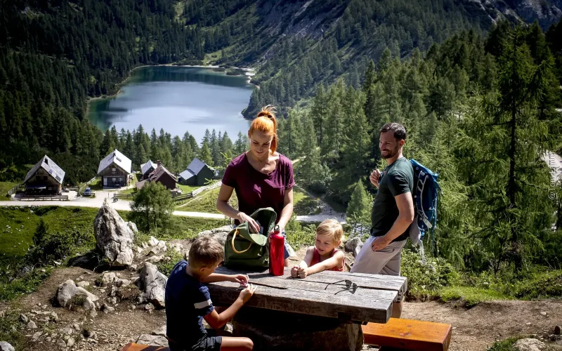 Familienrast beim Wandern am Rastplatz mit Blick auf Holzhütten im bewaldetem Talkessel und See im Hintergrund