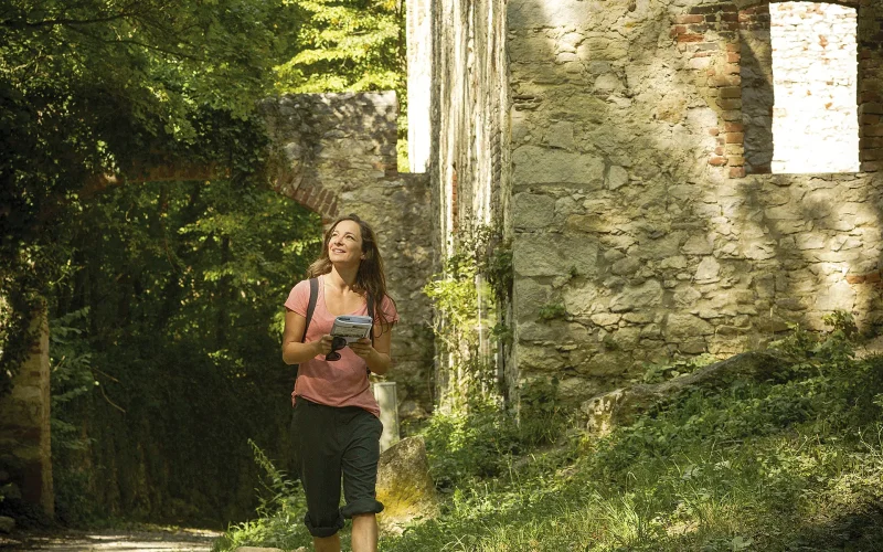 Mädchen in Wanderbekleidung mit Wanderkarte und Sonnenbrille in den Händen spaziert vor einer Steinruine auf einem gekiesten Wanderweg im Wald