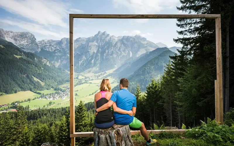 Rückansicht eines sich umarmenden, auf einem Holzstumpf sitzenden Pärchens in soomerlicher Sportbekleidung, mit Blick durch einen meschenhohen Holzrahmen in ein grünes Tal mit bewaldeten Hängen, einer Ortschaft in der Talsenke und einer Bergkette am Horizont